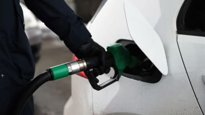 En vit personbil tankas vid en mack. Handen som håller i bensinpistorlen är klädd i en svart handske.