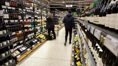 Alkon myymälän sisällä pulloja hyllyissä. Naismyyjä täyttää hyllyä ja asettaa juuri pulloja paikoilleen. Miesasiakas tummansinisessä toppatakissa ja musta pipo päässä kävelee ohi poispäin kamerasta. 