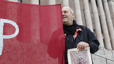 Kuvataiteilija Olavi Fellman seisoo Eduskuntatalon portailla ja pitää kiinni punaisesta banderolista.