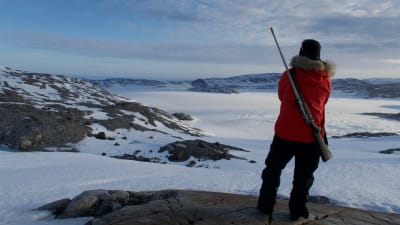 En person iklädd vinterkläder med pälskantad huva. Har ett gevär på ryggen. Står på ett berg och tittar ut över ett snölandskap. Befinner sig på Grönland. 