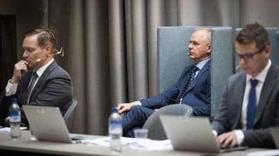 Finansinspektionens chef Tero Kurenmaa, tillbakalutad i en fåtölj mellan två kostymklädda herrar. 