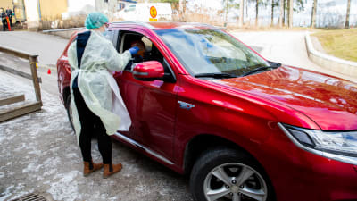 En sjukvårdare i skyddsutrustning ta ett coronavirustest på en person som sitter i en röd bil.