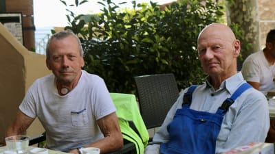 Två män sitter på kafé och ser in i kameran.