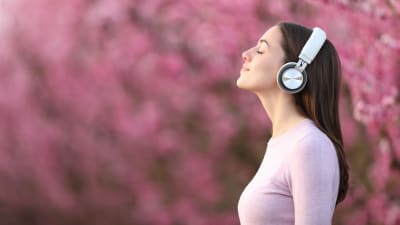 En kvinna med hörlurar på sig fotograferad från sidan. Hon står utomhus i ett fält av rosa blommor och träd.