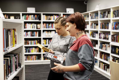 Harmaaseen collegeen pukeutunut ruskeahiuksinen nainen ja vankilavaatteisiin pukeutunut punahiuksinen nainen katsovat kirjoja kirjahyllyjen keskellä.