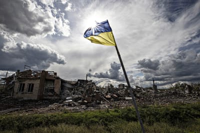 Den ukrainska flaggan vajar i motljus, i bakgrunden ett bostadshus i ruiner. 