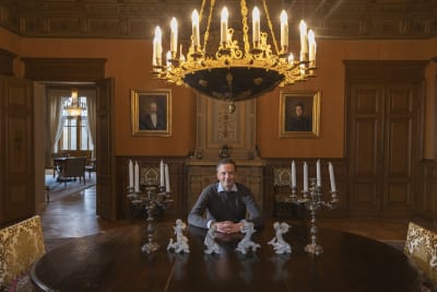 Henrik Creutz har händerna knäpta och sitter vid ett stort matbord. Takkronan hänger ovanför honom.