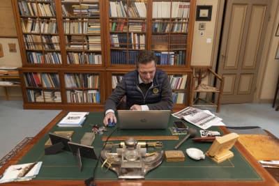 Henrik Creutz sitter vid ett skrivbord framför sin laptop, en stor bokhylla i bakgrunden.
