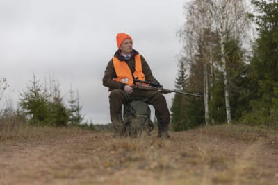 Henrik Creutz sitter på en pall mitt på sandvägen. Han har en orange väst. Han har ett gevär i handen.