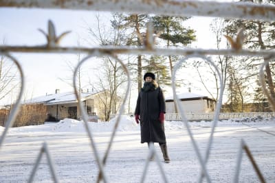En kvinna med lång svart vinterkappa fotograferad genom ett glest galler på en snöig väg, 