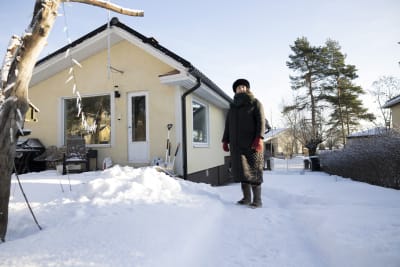 Kvinna i svart kappa och pälsmössa står på en snöig gård framför ett egnahemshus. 