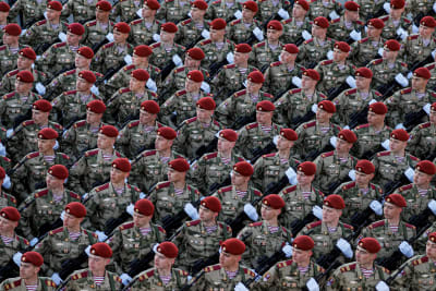 Suuri rivistö sotilaita marssii punaisissa bareteissa ja rynnäkkökiväät edessään ojennettuina katsoen vasemmalle.