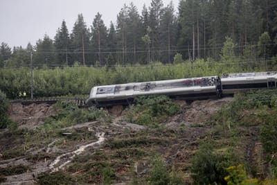 Ett tåg hänger från trasig räls i en sluttning som har rasat på grund av regnvatten. I bakgrunden syns tallskog.