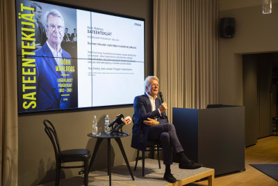 Björn Wahlroos vid bokpresentation, med mikrofoner, och bokbild på väggen bakom sig.