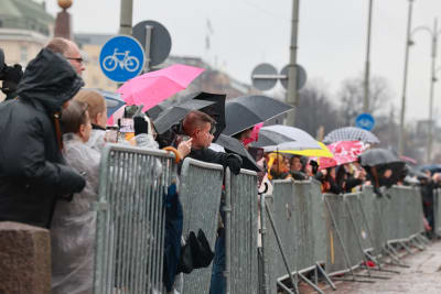 Folk med regnrockar och paraplyer väntar vid ett kravallstaket.
