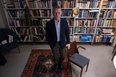 Henrik Meinander står och tittar upp mot kameran med ena handen på en stol. Bakom komom är väggen täckt av bokhyllor.