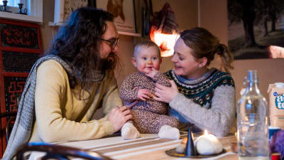 En åtta månader gammal bebis sitter på ett matbord och skrattar. Bredvid henne sitter en man och en kvinna.