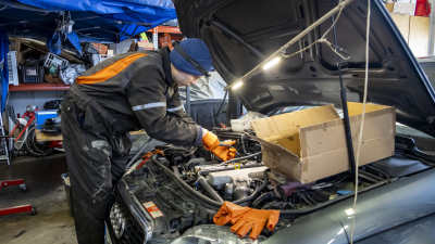 En ung man i svart overall, handskar och pannlampa står nedböjd över den öppna motorhuven på en bil i en bilverkstad.