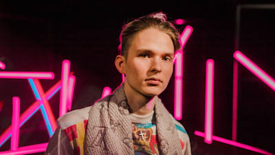 Vaaleahiuksinen nuori mies katsoo suoraan kameraan vakavalla ilmeellä pinkkien neonvalotolppien edessä.