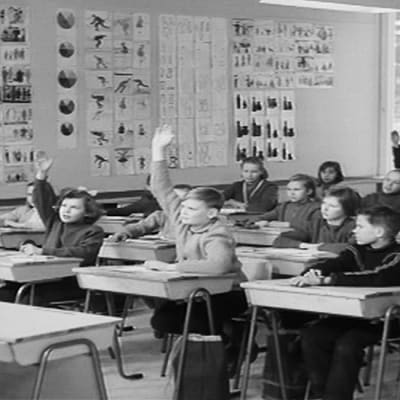 Oppilaita ja opettaja luokkahuoneessa, opettaja näyttää paperia ja oppilaista osa viittaa.