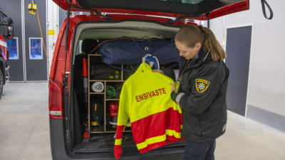 En kvinna står bakom en räddningsbil. Hon håller i en gul arbetsrock där det står "Ensivaste" på ryggen.