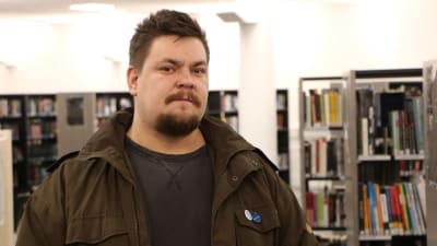 Mathias Rosenlund med brun jacka i ett bibliotek, tittande rakt in i kameran. 