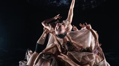Ljusklädda dansare mot en mörk bakgrund.