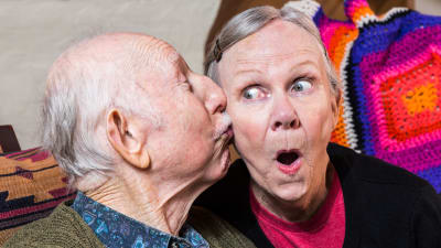 äldre man kysser äldre kvinna på kinden, hon ser förvånad ut