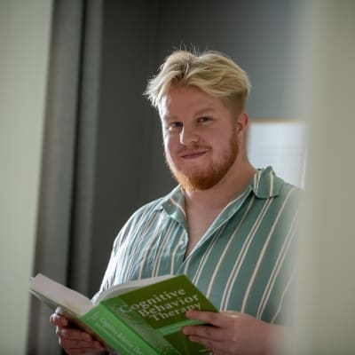 Sami-Petteri Asikainen pitelee kädessään vihreää oppikirjaa, jonka kannessa lukee Cognitive Behavior Therapy. Hän katsoo kameraan hymyillen.