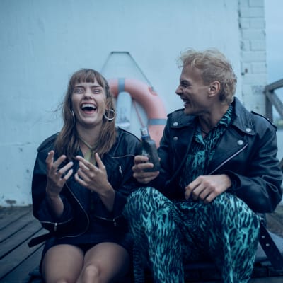 En ung tjej och en ung kille i läderjackor sitter skrattand på en brygga.