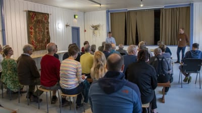 Många personer sitter samlade på bänkar och stolar under ett möte och lyssnar till en kvinna som talar.