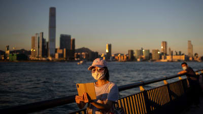Kvinna i Hongkong med ansiktsskydd tar en selfie med skyskrapor i bakgrunden. 