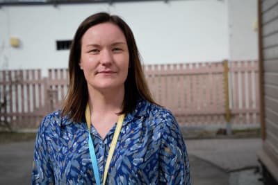 Brunhårig kvinna i blåspräcklig blus poserar vid en infart. 