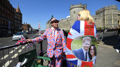 En man i kostym prydd med Storbritanniens flagga och en kvinna iklädd Storbritanniens flagga står framför Windsor slott.