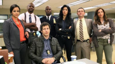 Ur serien Brooklyn Nine-Nine, skådespelarna Melissa Fumero, Terry Crews, Andy Samberg, Andre Braugher, Stephanie Beatriz, Joe Truglio Lo, Chelsea Peretti poserar i en gruppbild i en polisstation.