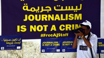 En man med kamera står framför en skylt där det står "journalistik är inte ett brott".