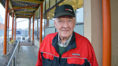 En äldre man i röd jacka står utanför en matbutik. Han tittar bestämt in i kameran.