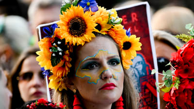 Kvinna ser allvarlig ut. Hon står i folkmassa iklädd en blomsterkrans med gula solrosor och blåa blommor. Hon har en ansiktsmålning som föreställer Ukraina.
