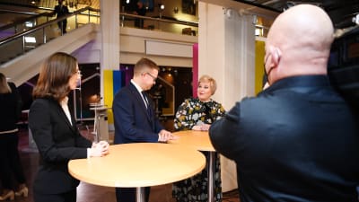 SDP:s ordförande Sanna Marin (till vänster), Samlingspartiets Petteri Orpo ( i mitten ) och Centerns Annika Saarikko (till höger) står vid höga bord och pratar med varandra.