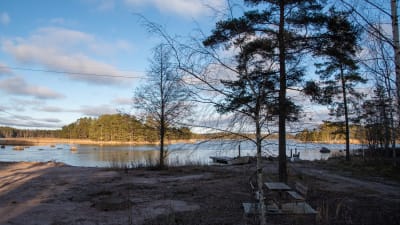 En bred strand. Till vänster sandstrand, till höger brygga och båt. Solen lyser på holmarna längre ut.