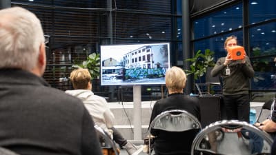 Några människor på stolar framför en scen och en tv som visar en bild på ett förfallet hus med graffiti. Bredvid xcenen står en man och talar i en orange kubformad mikrofon.