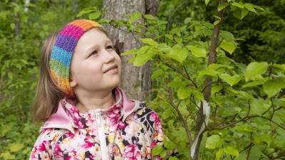 En sexårig flicka står i en träddunge, klädd i en blommig rock. Hon står och tittar på ett ungt träd.