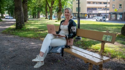 En ung kvinna sitter på en bänk i en park med en laptop i famnen. Hon tittar in i kameran och ler.