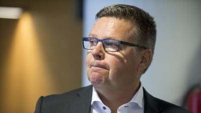 En medelålders man i kostym, skjorta och glasögon har en sammanbiten min.