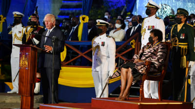 Prins Charles står vid ett podium och håller tal. På en stol på en upphöjning sitter Barbados president Sandra Mason och lyssnar. Omkring dem syns ett antal dignitärer i uniform.