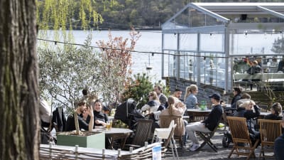 Människor på utomhusrestaurang i Stockholm med växthus och hav i bakgrunden. 