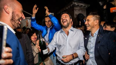 Matteo Salvini står med en Champagneflaska i handen och ser strålande glad ut.