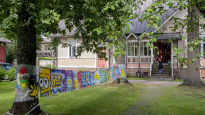 På bilden ser man ett konstverk hänga mellan träden. I bakgrunden ser man ett ljusrött hus och en människa gå upp för trappan.