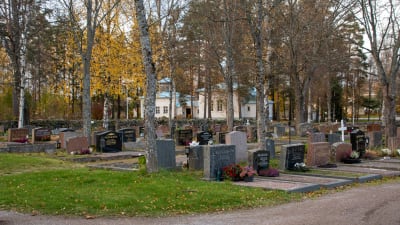 En begravningsplats. Gravstenar med blommor står i långa rader vid en grusväg. I bakgrunden syns träd i höstiga färger, en låg stenmur och ett vitt trähus. 