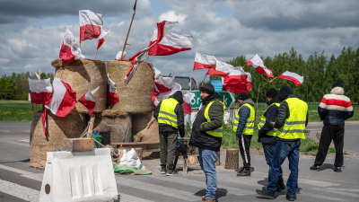 Några män i reflexväst står med händerna i fickorna framför fem höbalar med tiotals polska flaggor instuckna i sig.